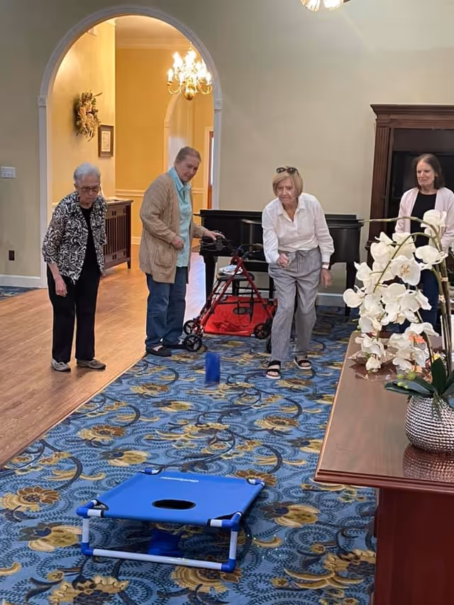 Four elderly women playing an indoor game of cornhole in a room with patterned blue carpet, wooden flooring, and a wooden table with a white orchid flower arrangement. One woman is tossing a bean bag towards a blue cornhole board on the floor. The room has a grand piano and a chandelier visible in the background.
