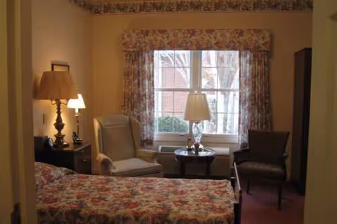 A bedroom with a floral bedspread, lamps, chairs, and a window with floral curtains.