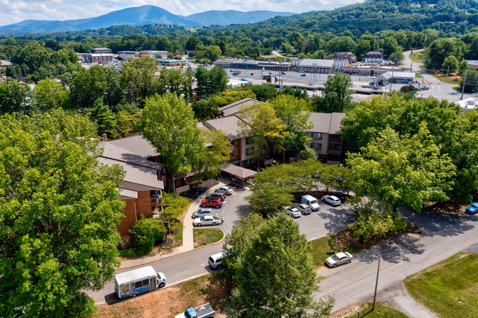 Aerial view of a multi-building senior living complex surrounded by trees with cars in the front driveway and mountains in the distance.