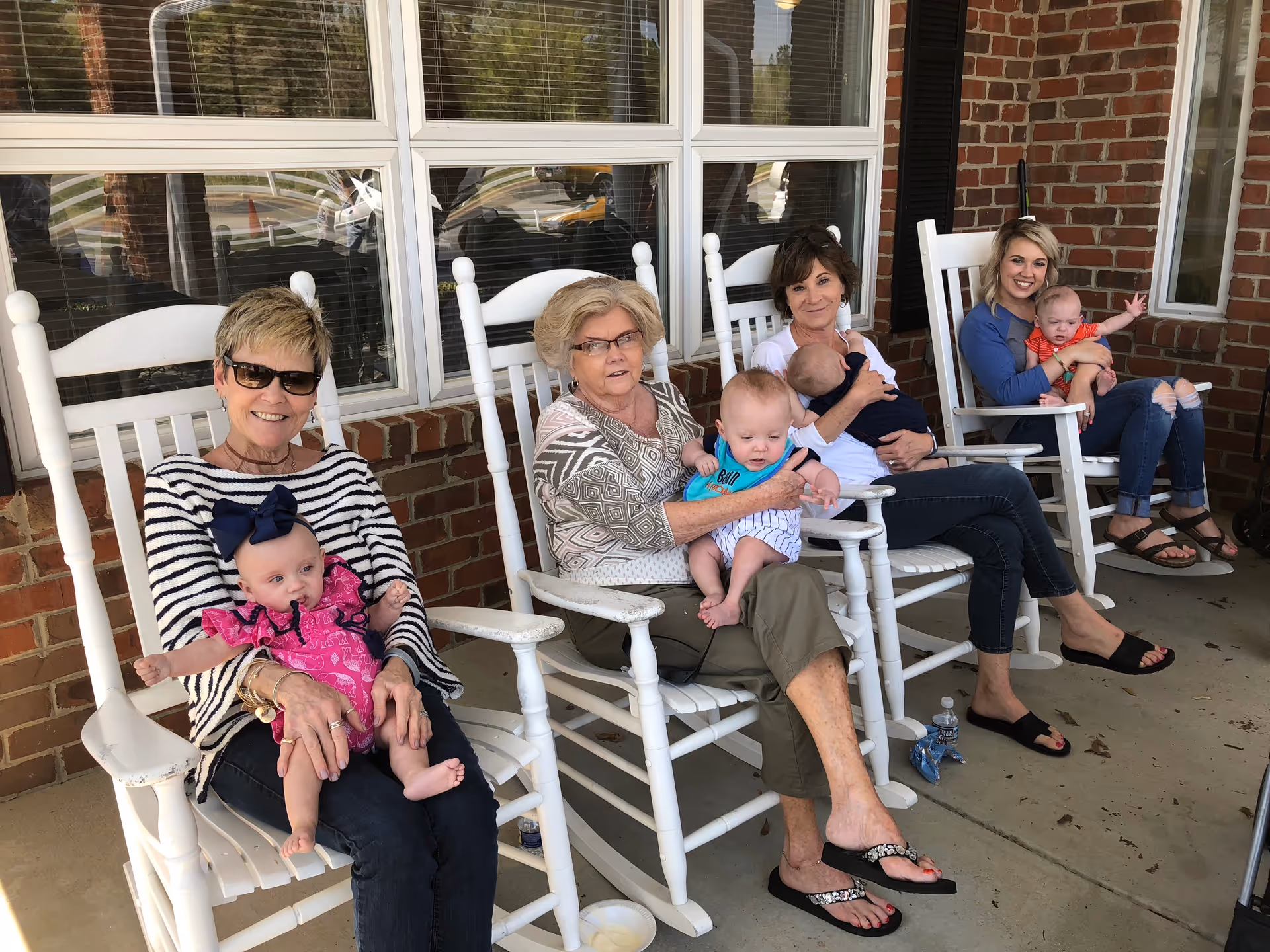 Four women sitting on white rocking chairs on a porch outside a brick building, each holding a baby. The women are smiling and appear to be enjoying a sunny day.