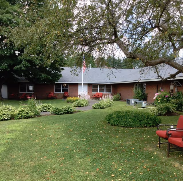 A single-story brick building with a gray roof surrounded by a well-maintained garden and lawn. There are several bushes and flowering plants, a flagpole with an American flag, and red outdoor chairs placed near the building and on the lawn. A large tree with overhanging branches is visible in the foreground.
