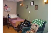 A small room with a single bed covered with a pink blanket, a wheelchair next to the bed, a green armchair with a snowflake-patterned blanket and a decorative pillow, a wooden nightstand with an American flag, and a green wall with a clock, framed picture, and two wall-mounted lights.