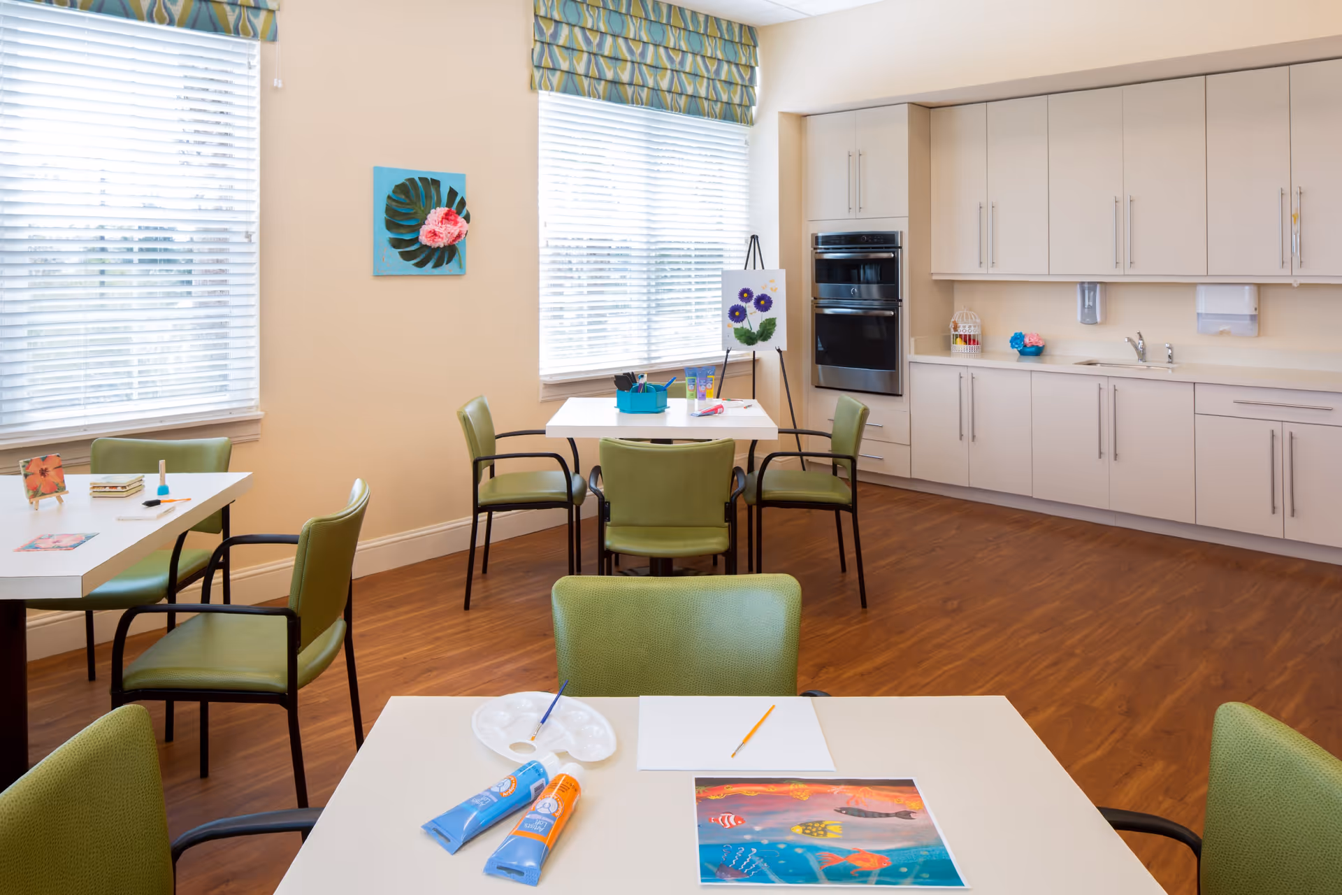 A bright and clean activity room with several tables and green chairs arranged around them. On the tables are art supplies including paint tubes, brushes, a palette, and a colorful painting. The room has large windows with blinds and patterned valances, light-colored walls, wood flooring, and a kitchenette area with cabinets, a sink, and built-in ovens. A small easel with a flower painting is also visible near the windows.