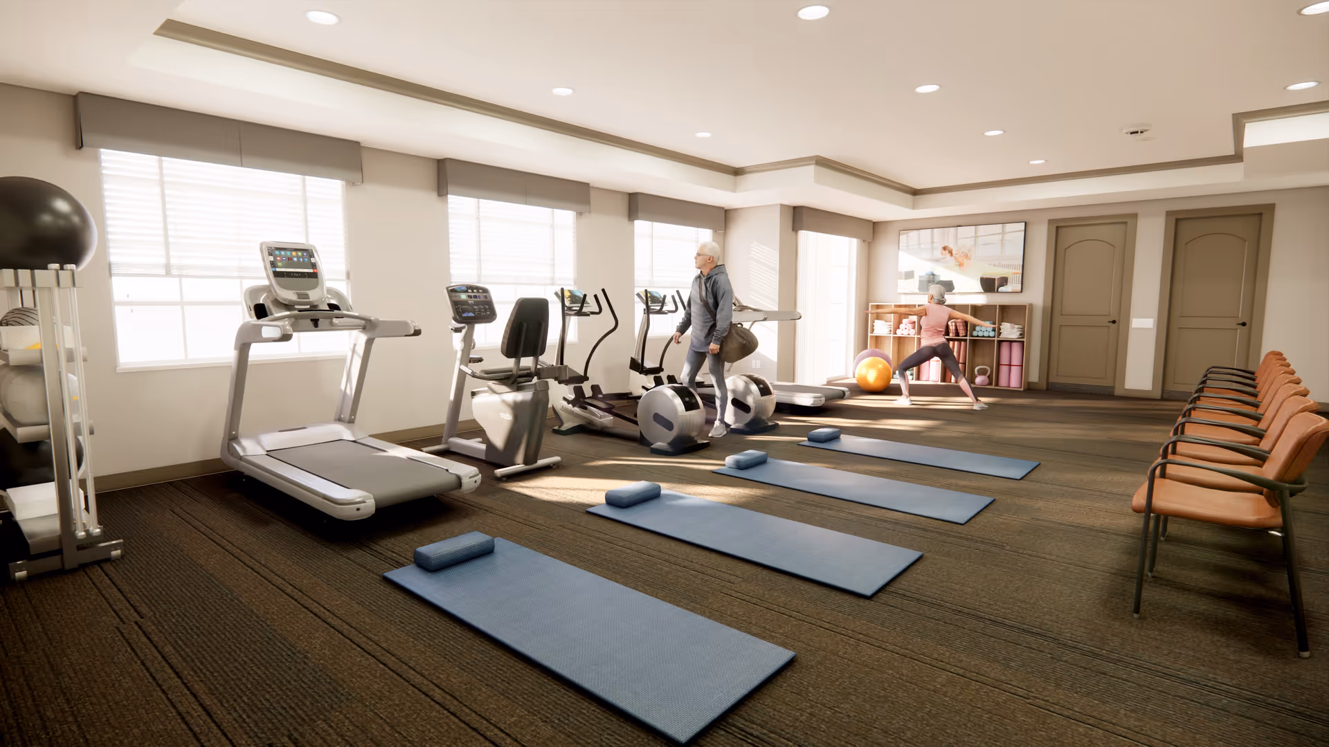 A bright fitness room with exercise equipment including treadmills, stationary bikes, and elliptical machines. Three blue exercise mats with small blue cushions are laid out on the carpeted floor. Two people are present: one older adult walking near the exercise machines and another person stretching near a shelf with exercise balls and yoga blocks. Several chairs are lined up against the wall on the right side. Large windows allow natural light to fill the room.
