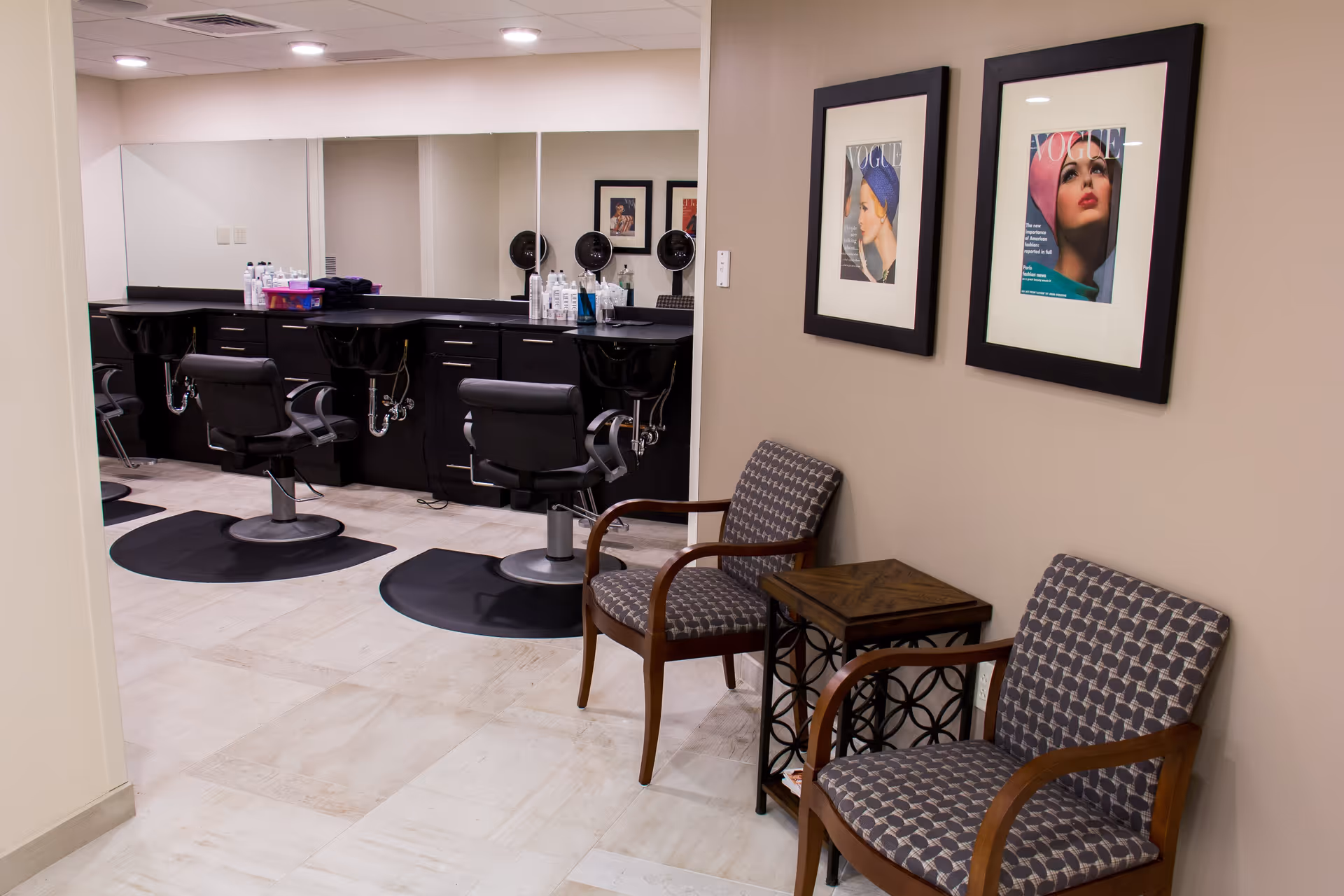 Interior view of a salon area with two black salon chairs in front of a counter with sinks and hair care products. There are two patterned armchairs with wooden arms and a small wooden side table between them. Two framed Vogue magazine covers are hanging on the beige wall above the chairs.