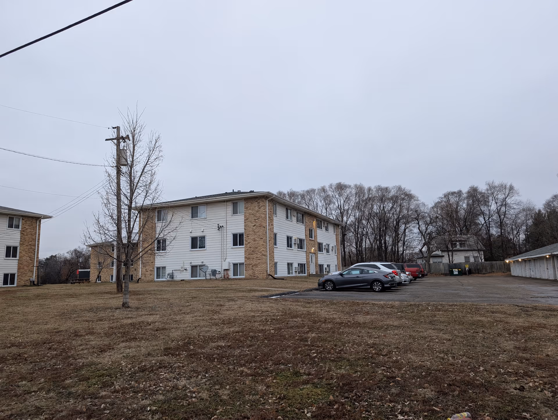 Exterior view of a three-story residential building with brick and white siding, surrounded by a grassy area and a parking lot with several parked cars. Leafless trees and a cloudy sky are visible in the background.