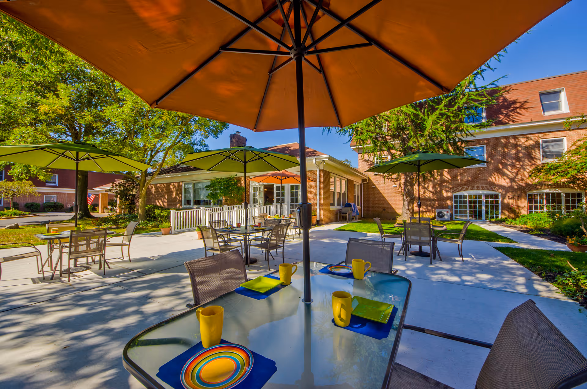 Outdoor patio area with several tables and chairs under large umbrellas. Each table is set with colorful plates, napkins, and yellow mugs. The patio is surrounded by greenery and trees, with a brick building in the background under a clear blue sky.