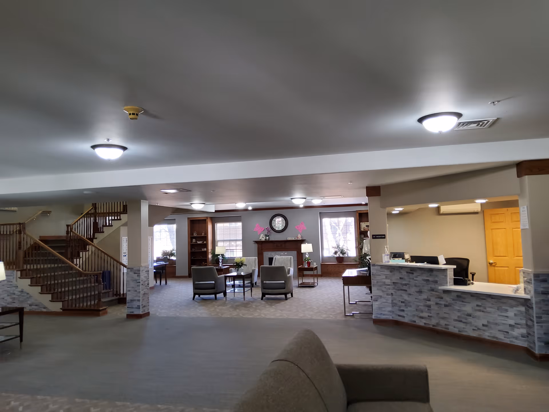 Spacious assisted living facility common area with seating, a reception desk, a fireplace, and a staircase.