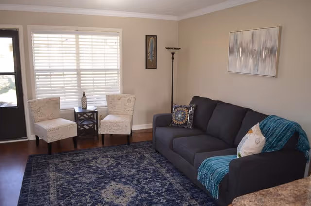 A cozy living room with a dark gray sofa adorned with decorative pillows and a teal throw blanket. Two light-colored patterned chairs are positioned near a window with white blinds, separated by a small dark wooden table holding a decorative item. A tall floor lamp stands in the corner, and a framed abstract painting hangs on the beige wall above the sofa. The floor is covered with a large blue patterned rug.
