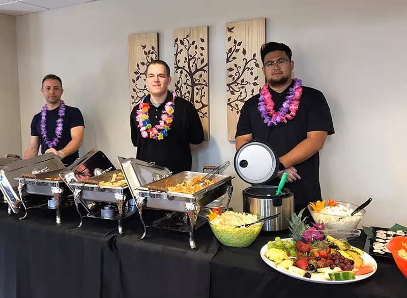 Three men wearing black shirts and colorful leis stand behind a buffet table with chafing dishes filled with food, bowls of salad, and a platter of fresh fruit in a room with decorative wooden wall art.