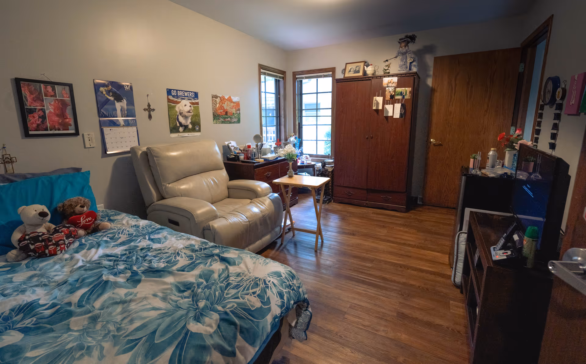 A cozy bedroom featuring a bed with blue floral bedding and two teddy bears, a beige recliner chair, a small wooden table with a vase of flowers, a wooden dresser, a wooden wardrobe with photos and decorations on it, and a TV on a stand. The room has wooden flooring, two windows letting in natural light, and various wall decorations including a calendar and pictures.