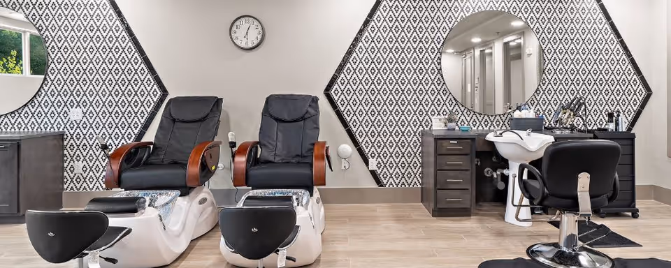 Interior view of a salon area in a senior living facility featuring two black leather pedicure chairs with foot baths, a black salon chair in front of a white sink, and a vanity with a large round mirror. The walls have a decorative black and white diamond pattern behind the chairs and mirror.