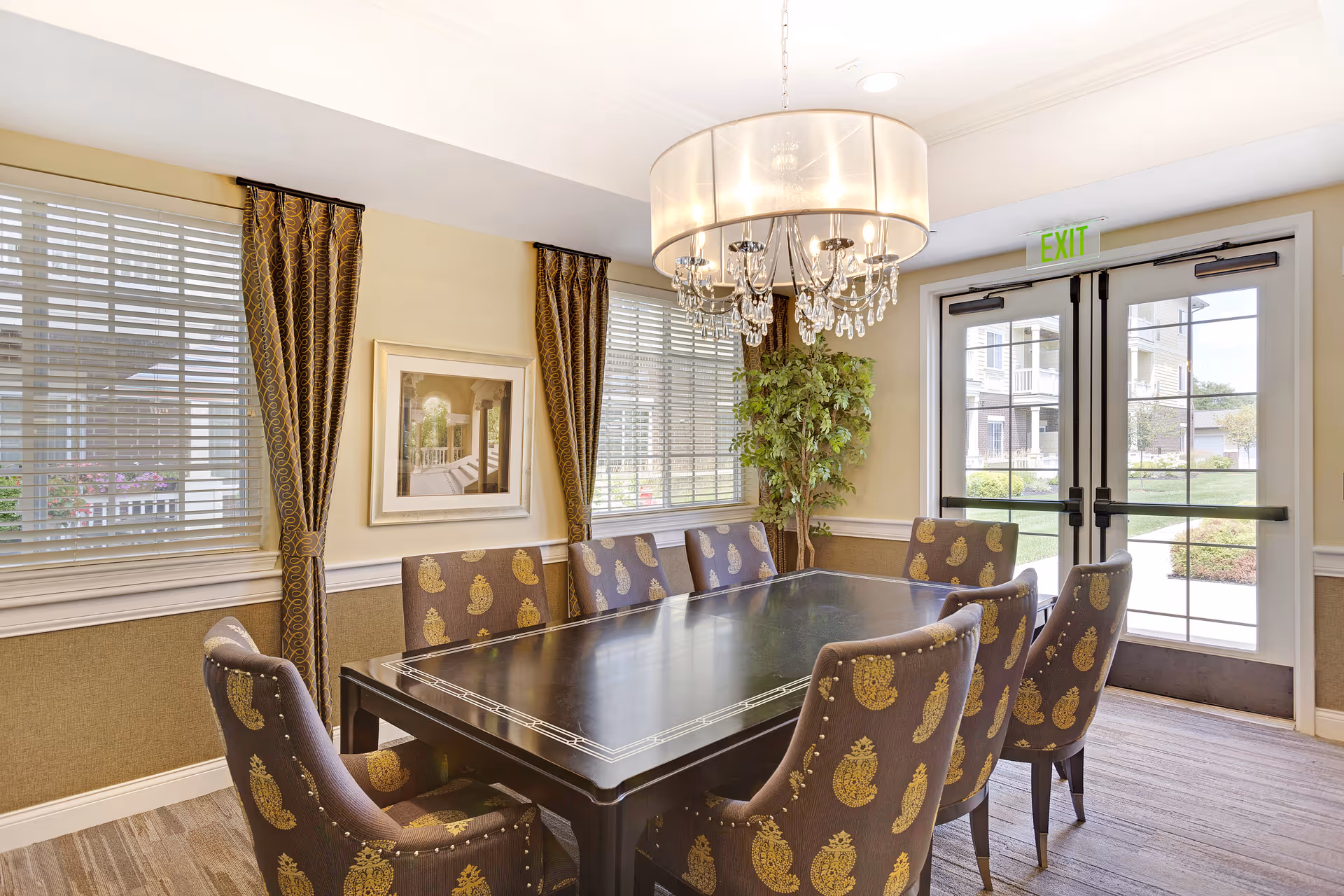 A well-lit dining room with a large rectangular dark wood table surrounded by eight upholstered chairs featuring a gold paisley pattern. The room has two large windows with white blinds and brown patterned curtains, a framed picture on the wall, a green potted plant in the corner, and a modern chandelier with crystal accents hanging above the table. Double glass doors with an exit sign lead outside to a landscaped area.
