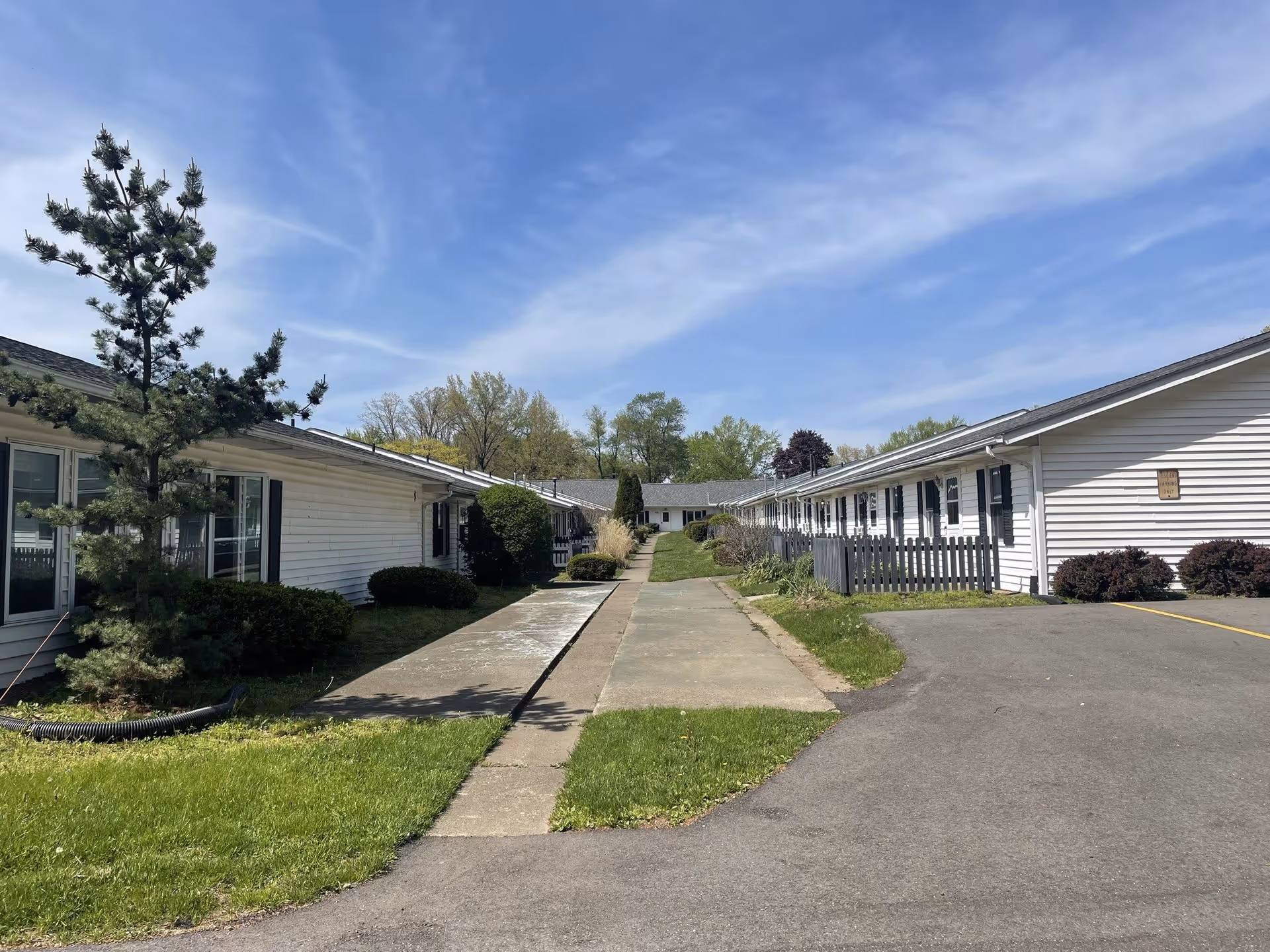 Exterior view of Glenwood Assisted Living facility showing single-story white buildings with black shutters on either side of a paved walkway. There are green lawns, bushes, and trees under a blue sky with some clouds.