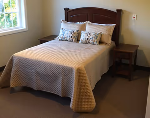 A neatly made bed with a beige quilted bedspread and four pillows, two of which have a leaf pattern. The bed has a dark wooden headboard and is flanked by two matching wooden nightstands. A window on the left side lets in natural light, and the walls are painted a light color.