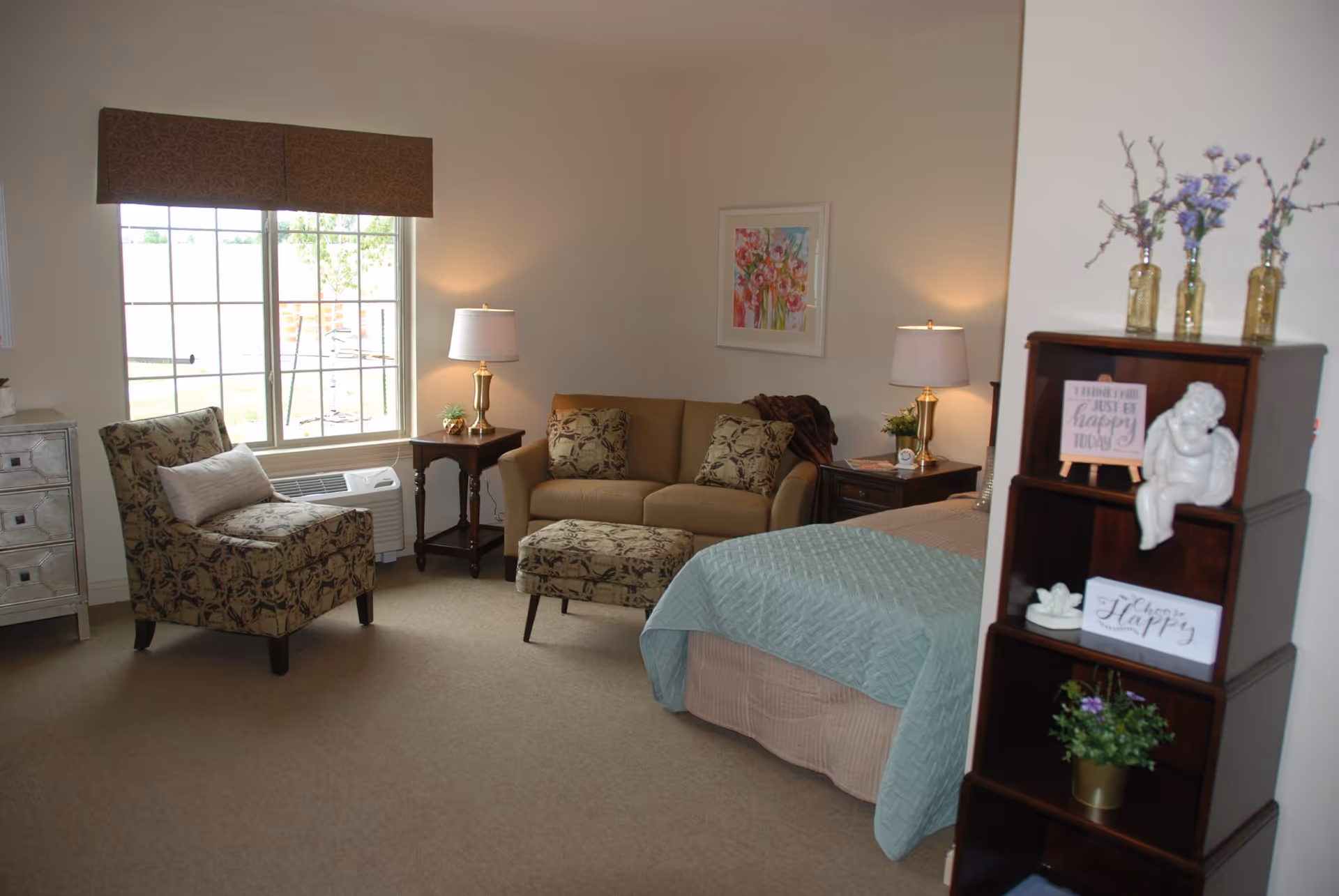 Light-filled senior living bedroom suite with a bed, loveseat, patterned armchair, ottoman, side tables and shelving.