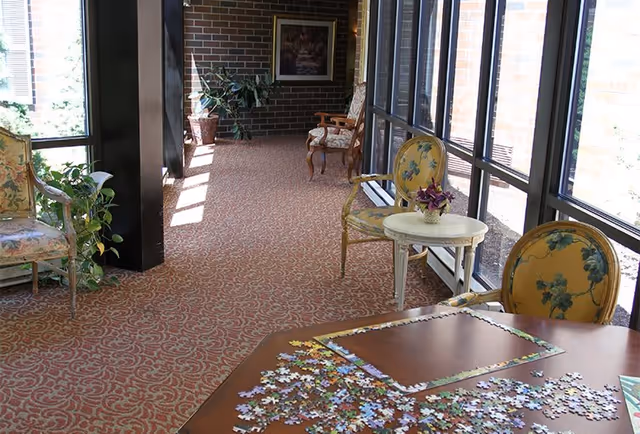 A cozy indoor lounge area with patterned carpet, comfortable chairs, and a table with a puzzle. Large windows allow natural light to fill the space, complemented by potted plants and artwork on the walls.