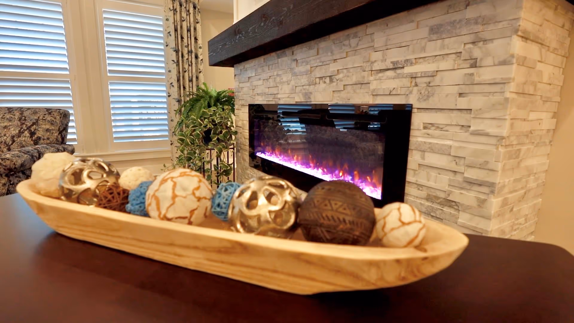A cozy living room area featuring a modern electric fireplace with a stone surround. In front of the fireplace is a wooden tray holding decorative spheres of various colors and textures. To the left, there is a patterned armchair and a window with white plantation shutters and floral curtains. A green potted plant is also visible near the fireplace.