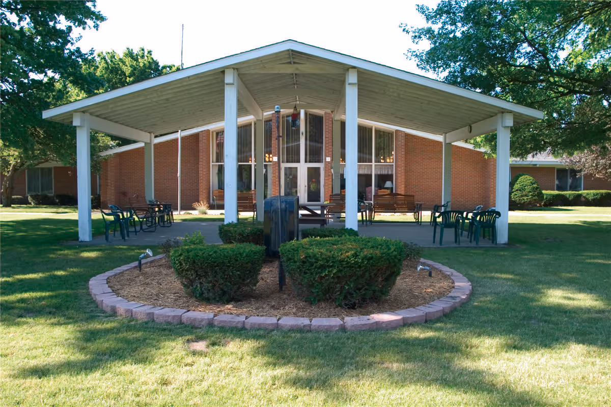 Covered outdoor pavilion with chairs and shrubs in front of a single-story brick senior living building.