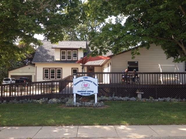Exterior view of Dansville Country Care assisted living facility showing a two-story building with beige siding and dark brown trim. There is a wooden ramp and deck in front with two people sitting on the deck under a red umbrella. The area is shaded by large trees and there is a sign in front of the building with the facility name and contact information.