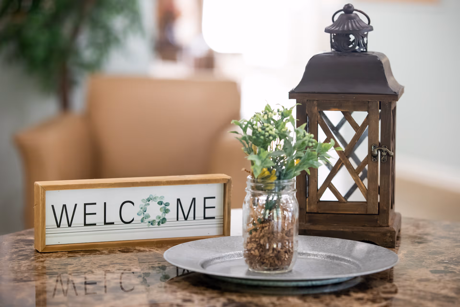 Tabletop welcome display with a 'WELCOME' sign, a small jarred plant, and a decorative lantern on a tray in a seating area.