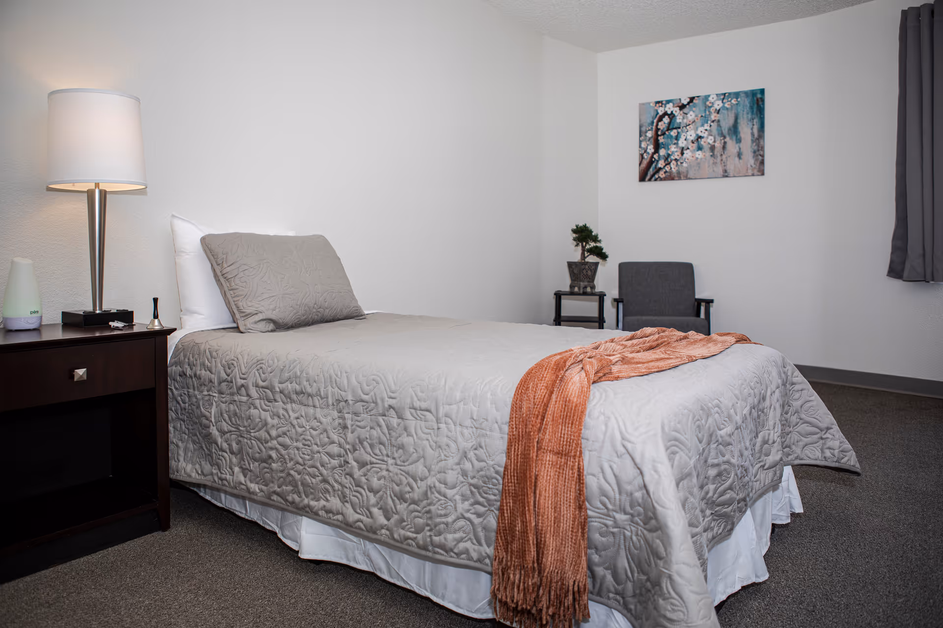 Neatly made single bed in a minimally decorated bedroom with a nightstand, lamp, chair, and wall art.