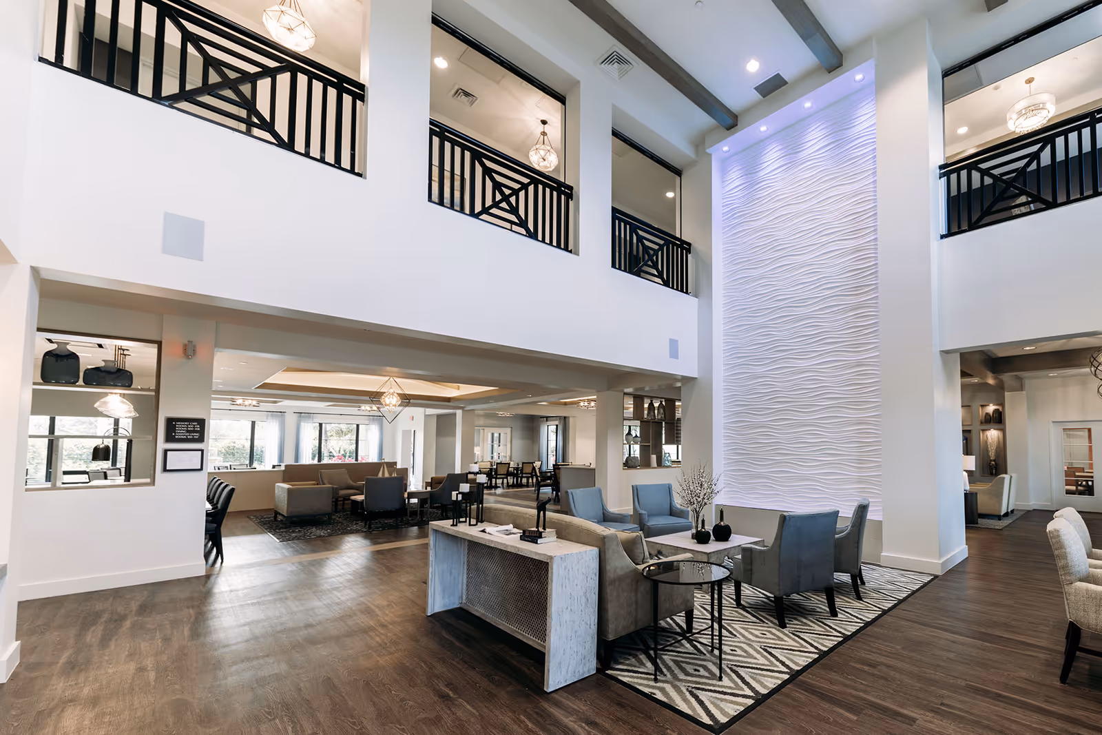 Spacious and modern senior living facility common area with high ceilings, dark wood flooring, and contemporary furniture including sofas and armchairs arranged around a textured wall with soft lighting. The area features large windows, decorative light fixtures, and an open layout with a second-floor balcony overlooking the space.