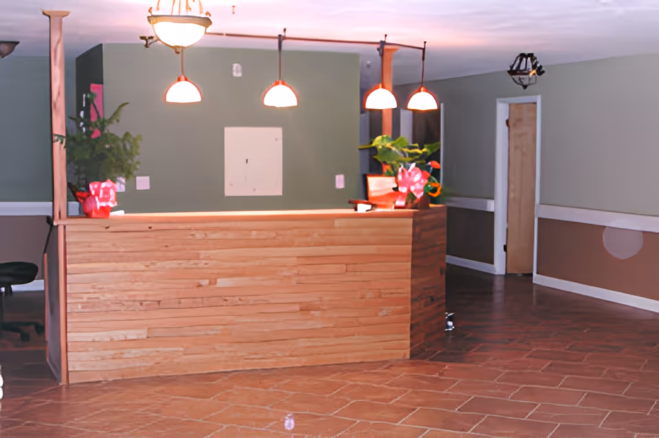 Reception area with a wooden front desk, three hanging pendant lights, a ceiling light fixture, potted plants with red wrapping on the desk, and a hallway with a door in the background.