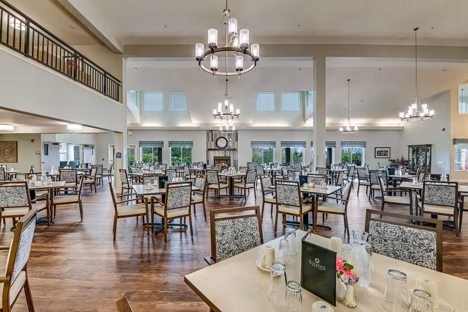 A spacious dining room in a senior living facility with multiple tables and chairs arranged neatly. The room features large windows allowing natural light, chandeliers hanging from the ceiling, and a fireplace with a clock above it. Tables are set with glasses, napkins, and menus labeled 'Solstice'.