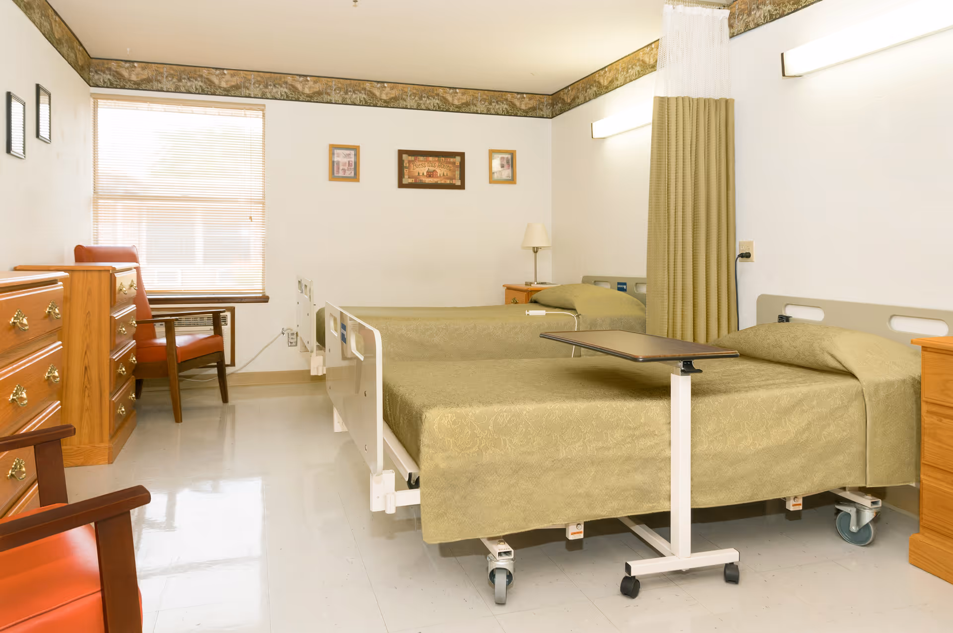 A clean and simple room in a care center with two hospital-style beds covered in green bedspreads. Each bed has a small wooden nightstand beside it, and there is a rolling overbed table positioned over one of the beds. The room has a window with blinds, two wooden dressers, and two wooden chairs with red cushions. The walls are white with a decorative border near the ceiling, and three framed pictures hang on the wall above the beds.