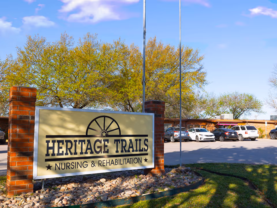Outdoor view of Heritage Trails Nursing & Rehabilitation facility sign with brick pillars, trees with green leaves, parked cars, and a clear blue sky.