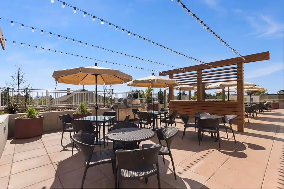 Outdoor patio area with multiple round tables and black wicker chairs, beige umbrellas providing shade, string lights overhead, and a barbecue grill against a railing with plants in large pots under a clear blue sky.