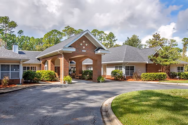 Exterior view of a single-story senior living facility building with a covered entrance supported by brick columns, surrounded by greenery and trees under a partly cloudy sky.