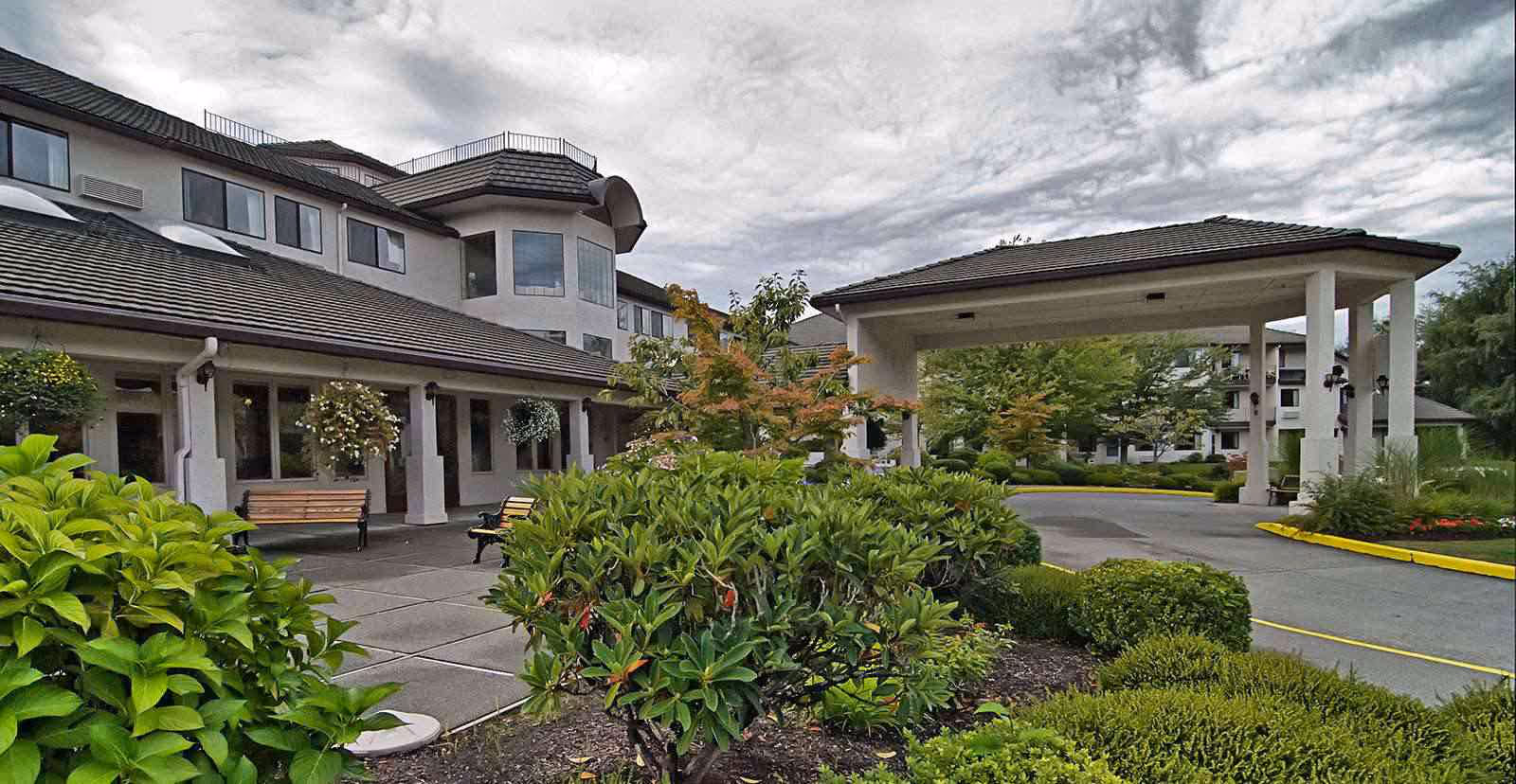 Exterior front entrance of a multi-story senior living building with a covered porte-cochere, benches, and landscaped shrubs.