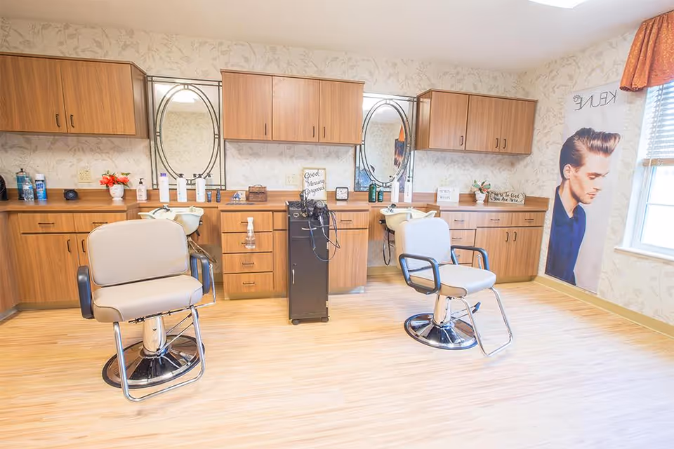A bright and clean hair salon area with two beige salon chairs in front of wooden cabinets and two oval mirrors. The room has light wood flooring, floral wallpaper, and a large poster of a man with styled hair near a window with blinds.