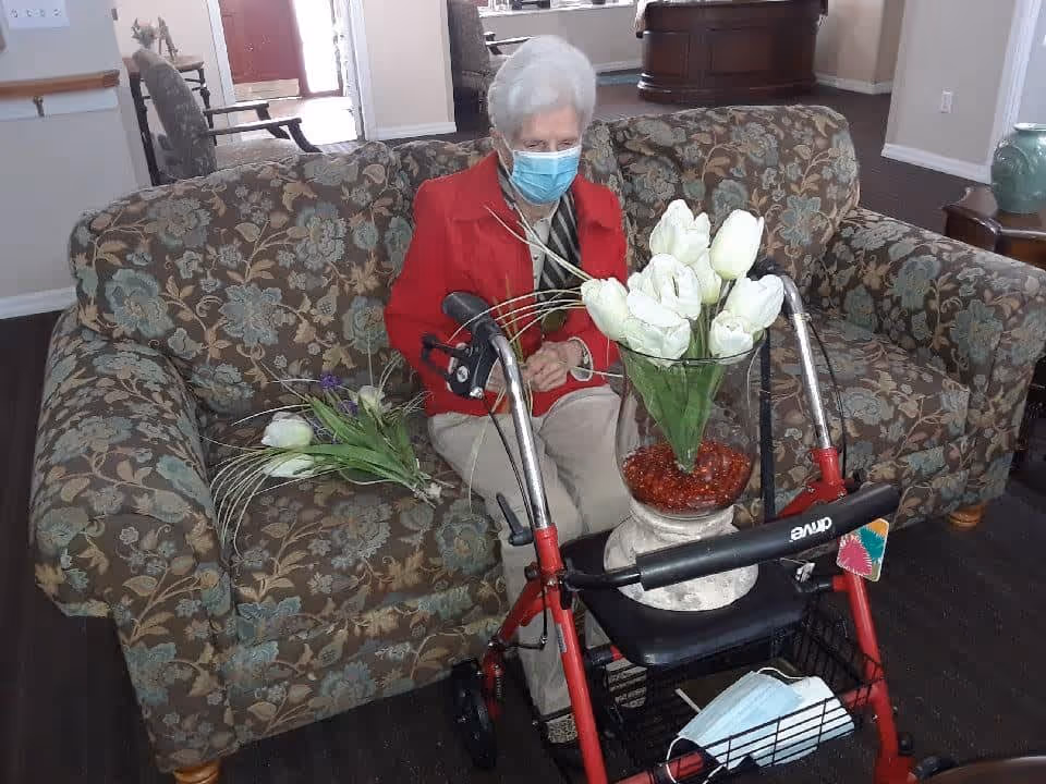 An elderly woman wearing a red jacket and a face mask sits on a floral patterned couch in a living room. In front of her is a red walker with a vase of white tulips placed on its seat. Another bunch of flowers lies on the couch beside her. The room has dark wood flooring and light-colored walls.