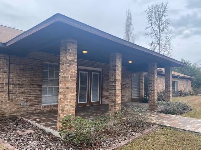 Exterior view of a single-story brick building with a covered porch supported by brick columns. The porch has two ceiling lights and a double glass door entrance. There are windows with blinds on either side of the door and some landscaping with bushes and grass in front.