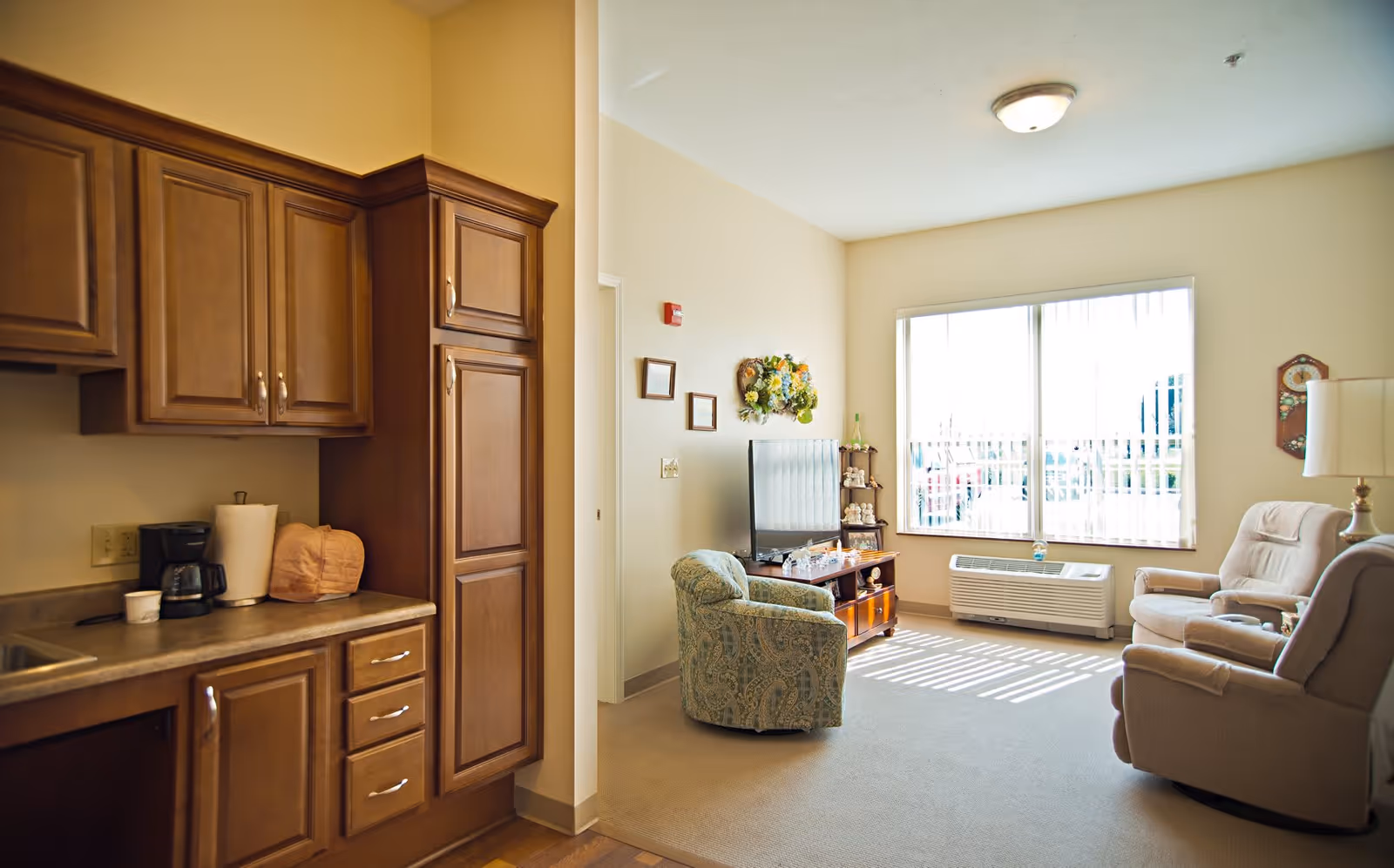 Interior view of a senior living facility room showing a small kitchenette area with wooden cabinets, a coffee maker, and paper towels on the counter. Adjacent to the kitchenette is a cozy sitting area with a patterned armchair, two beige recliners, a wooden TV stand with a flat-screen television, and a large window with vertical blinds letting in natural light.