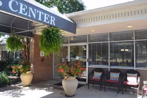 Entrance area of a facility with a covered awning labeled 'CENTER'. There are large potted plants with green foliage and flowers near the entrance. To the right, there are three black chairs with colorful cushions placed against a window with horizontal blinds.