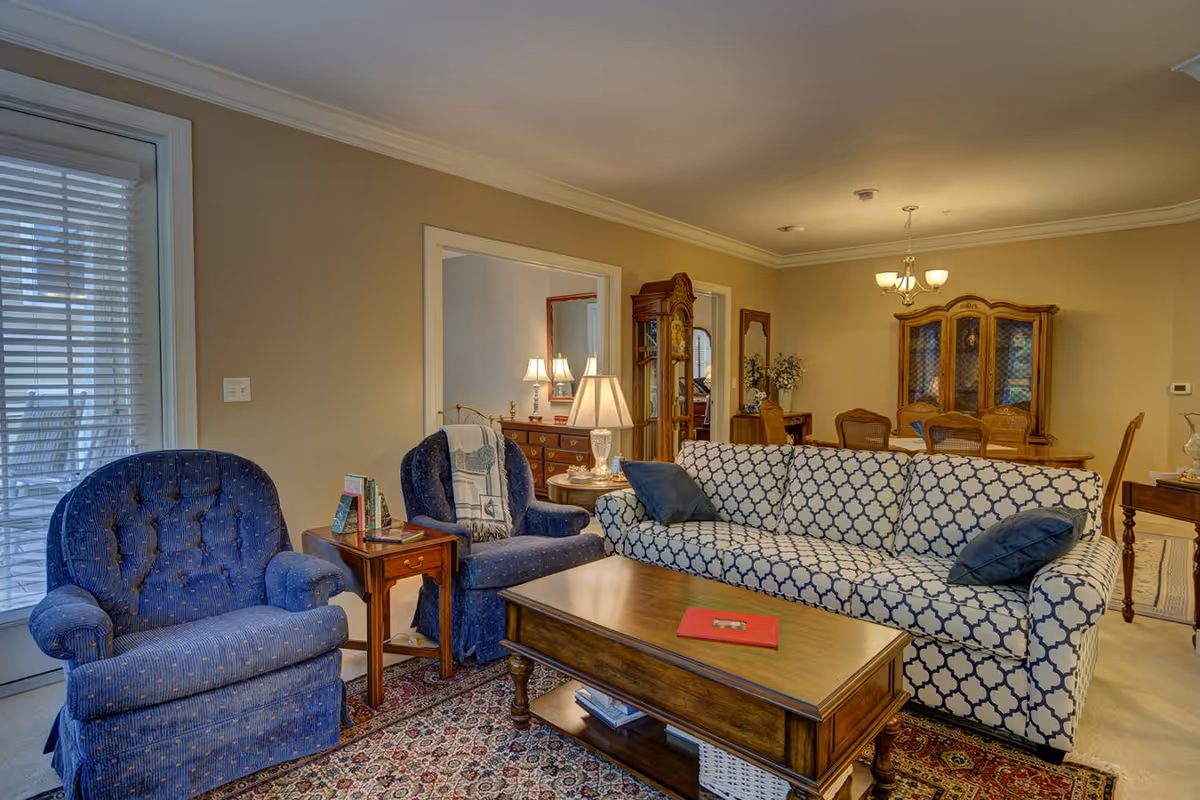 A cozy living room with a patterned blue and white sofa, two blue armchairs, a wooden coffee table with a red book on it, and a patterned rug. In the background, there is a dining area with a wooden dining table, chairs, and a china cabinet. The walls are painted beige and there are lamps providing warm lighting.