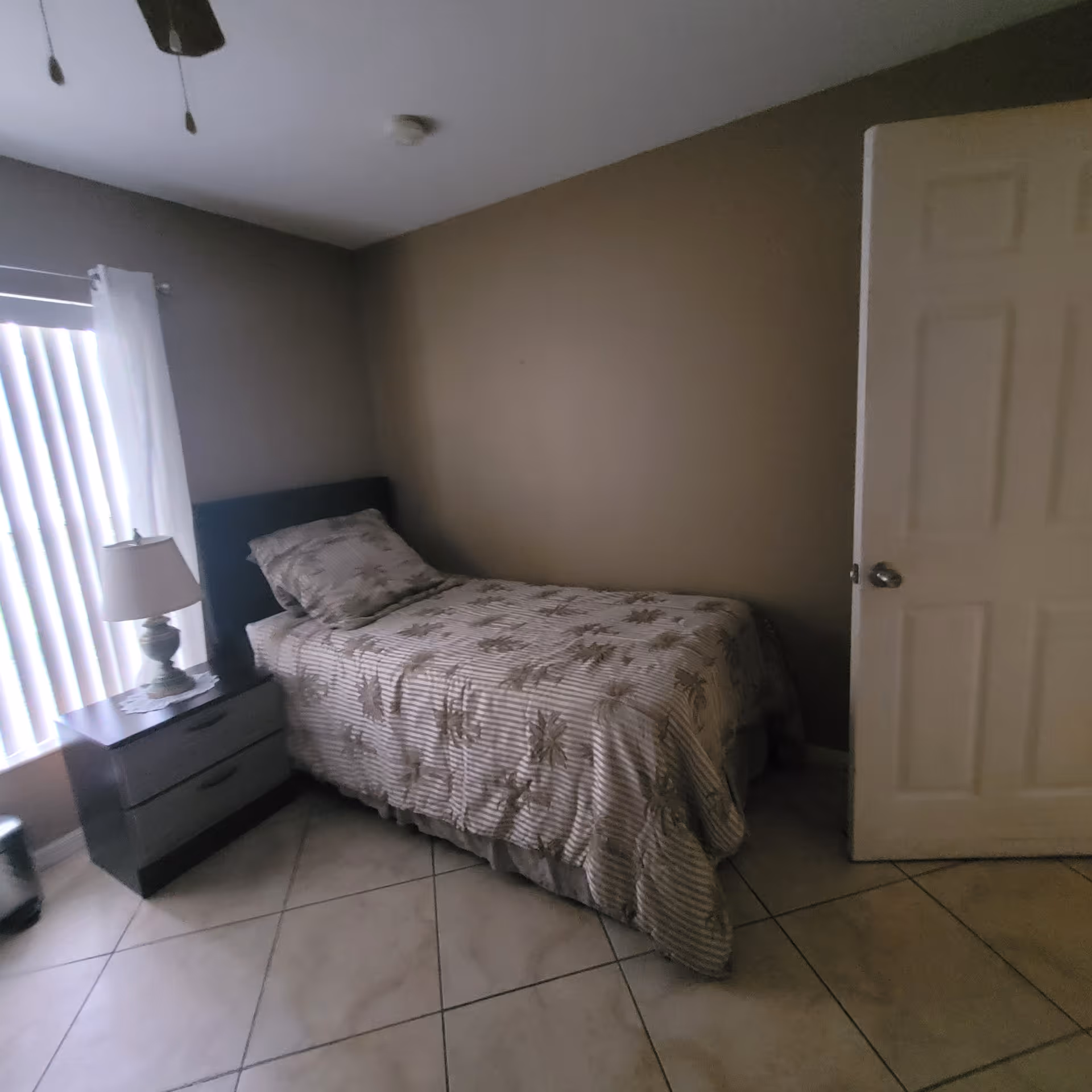 Small bedroom with a twin bed, nightstand and lamp beside a window with vertical blinds.