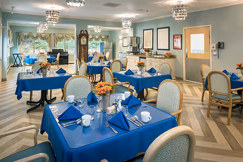 A bright dining room with multiple tables covered in blue tablecloths, each set with blue napkins, white cups, silverware, and small flower arrangements. The room features light blue walls, large windows with decorative curtains, a grandfather clock, and modern ceiling lights. Chairs with light upholstery surround the tables.