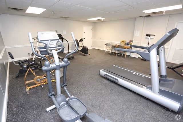 Indoor exercise room with various fitness equipment including a treadmill, elliptical machine, stationary bike, and a rowing machine. There is a table with chairs and some items on it against the far wall. The room has white walls and a gray rubber floor.