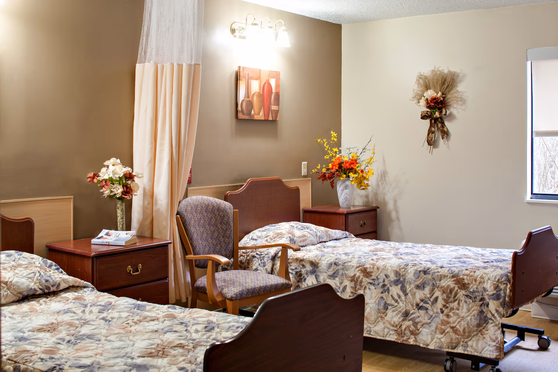 Two twin beds with floral bedspreads in a furnished senior living bedroom with nightstands, chairs, and decorative flowers.