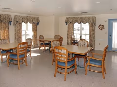 Dining room with several wooden tables and upholstered chairs, windows with patterned valances, and a ship wheel on the wall.