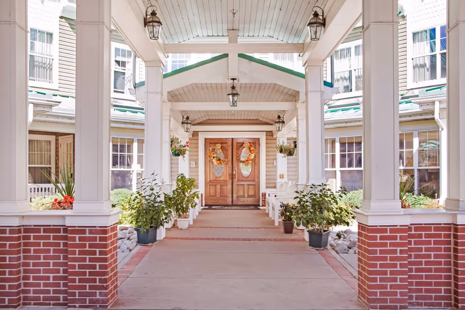 Covered entrance walkway leading to double wooden doors decorated with autumn wreaths, flanked by white columns with brick bases and potted plants on either side.