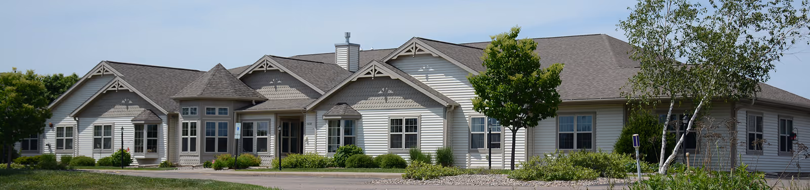 Single-story beige assisted living building with gabled roofs, landscaped lawn and trees under a clear sky.