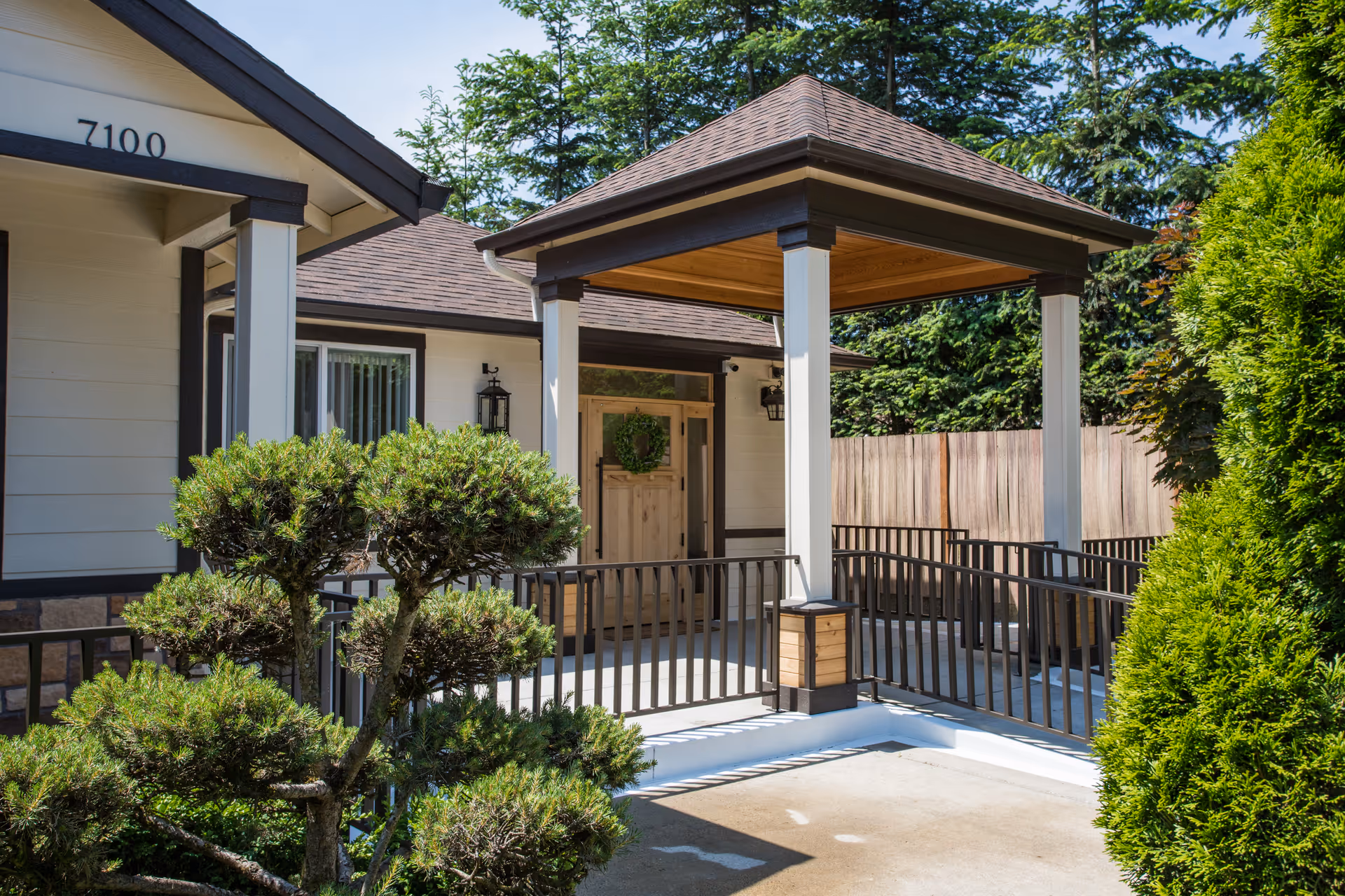 Entrance of a single-story building with a covered porch supported by white columns and a wooden door decorated with a green wreath. There is a ramp with black railings leading to the door, surrounded by well-maintained greenery and trees.