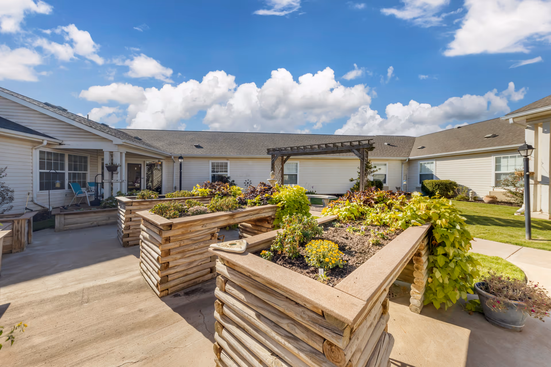 Courtyard with raised wooden garden beds and a pergola surrounded by single-story facility buildings under a blue sky.