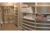 A spacious walk-in closet with multiple shelves and hanging rods. The shelves are neatly stocked with folded towels and linens in white and beige colors. The closet has a beige carpeted floor and light-colored walls.